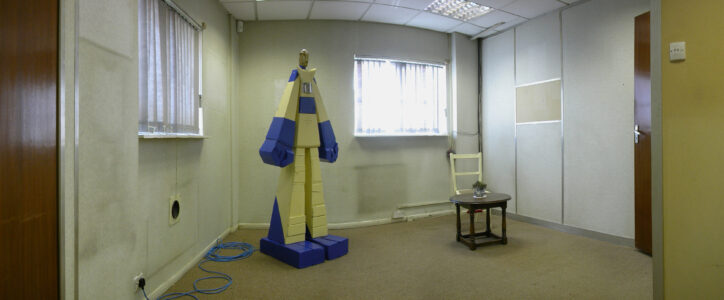 Art installation of a blue and yellow wooden robot a chair and a table in a room