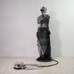 Contemporary sculpture of a female figure, similar to the "Venus de Milo", made of black fridge coils and loudspeakers connected to stones on the floor.